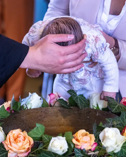 Bebê de vestido florido recebendo a bênção sobre a pia batismal enfeitada com rosas