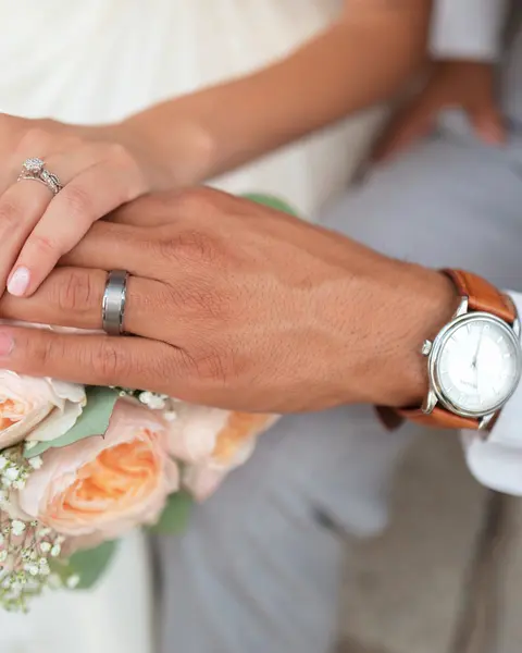 Mãos de um casal com alianças, sobre um buquê de rosas